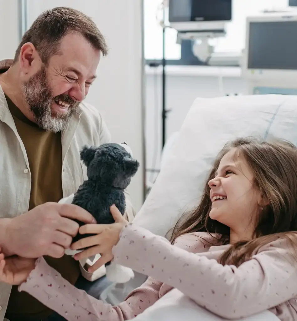 Das Bild zeigt ein Mädchen, dass in einem Krankenhausbett liegt. Neben ihr sitzt ein Mann - ihr Vater. Er gibt seiner Tochter ein Kuscheltier. Beide lachen. Das Bild wird im Bereich "Mitaufnahme der Eltern beim Krankenhausaufenthalt eines Kindes" verwendet.