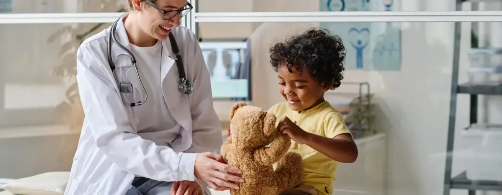Das Bild zeigt eine Ärztin mit kurzen Haaren, die einen weißen Arztkittel trägt und um den Nacken ein Stethoskop hängen hat. Sie sitzt auf einer Behandlungsliege neben einem Kleinkind, das einen Teddybären in den Händen hält. Beide Personen lachen. Das Bild wird im Bereich "Molekulardiagnostik Pädiatrischer Tumore" verwendet.