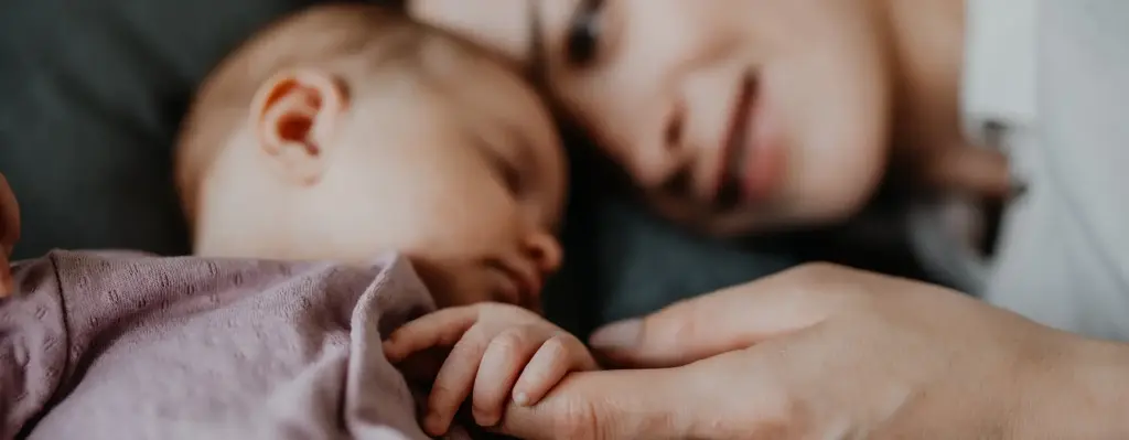 Das Bild zeigt eine Mutter, die mit dem Kopf an ihrem schlafenden Baby liegt. Scharf im Vordergrund sieht man wie das Baby den Finger der Frau hält. Das Bild wird im Bereich "Mutterschaft – Sicherheit durch unsere Unterstützung" verwendet.