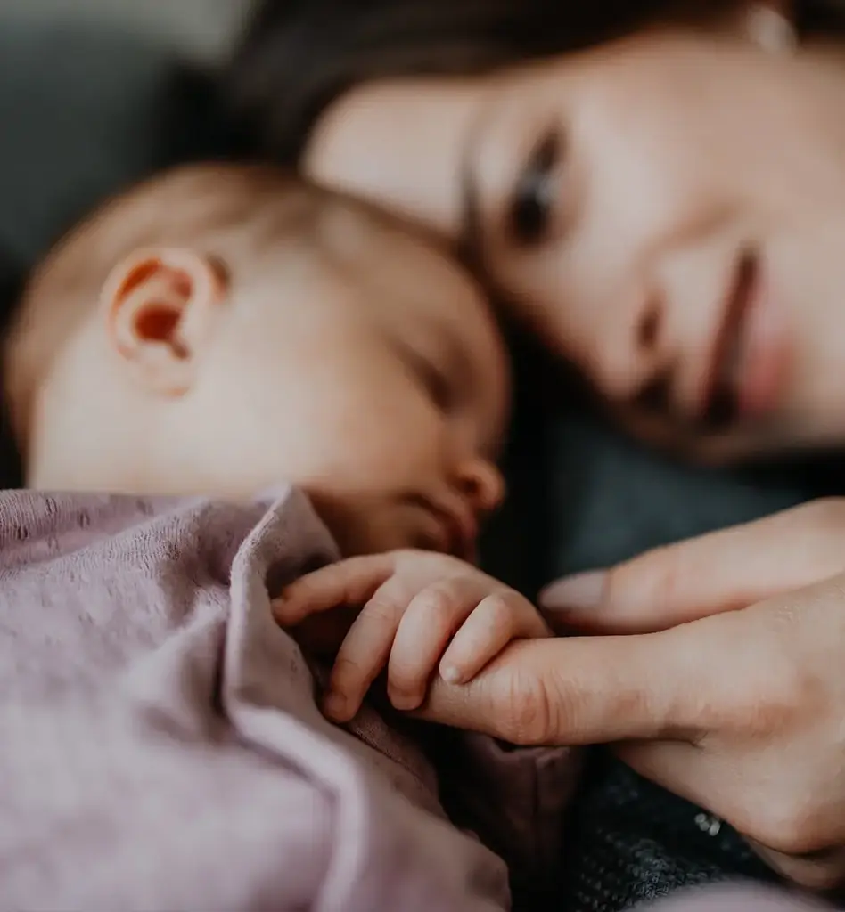 Das Bild zeigt eine Mutter, die mit dem Kopf an ihrem schlafenden Baby liegt. Scharf im Vordergrund sieht man wie das Baby den Finger der Frau hält. Das Bild wird im Bereich "Mutterschaft – Sicherheit durch unsere Unterstützung" verwendet.