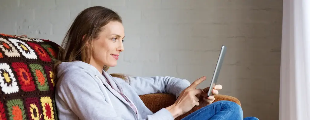 Eine Frau sitzt auf einem Sofa. Man sieht sie seitlich. Sie hält in der linken Hand ein Tablet und blickt auf den Bildschirm. Das Bild wird im Bereich "myReha – Individuelle Neuro-Therapie nach Schlaganfall und Schädel-Hirn-Trauma" verwendet.