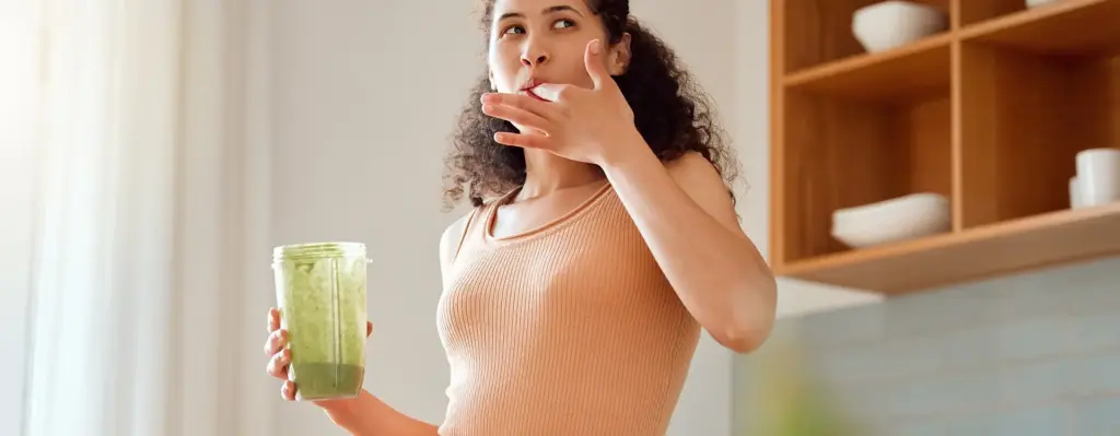 Eine Frau steht in einer Küche. Sie lutscht sich den Zeigefinger ihrer linken Hand ab. In der rechten Hand hält sie einen Behälter für einen Mixer, in dem ein grüner Smoothie drin ist. Das Bild wird im Bereich "myWEIGHTcoach und myHEALTHcoach" verwendet.