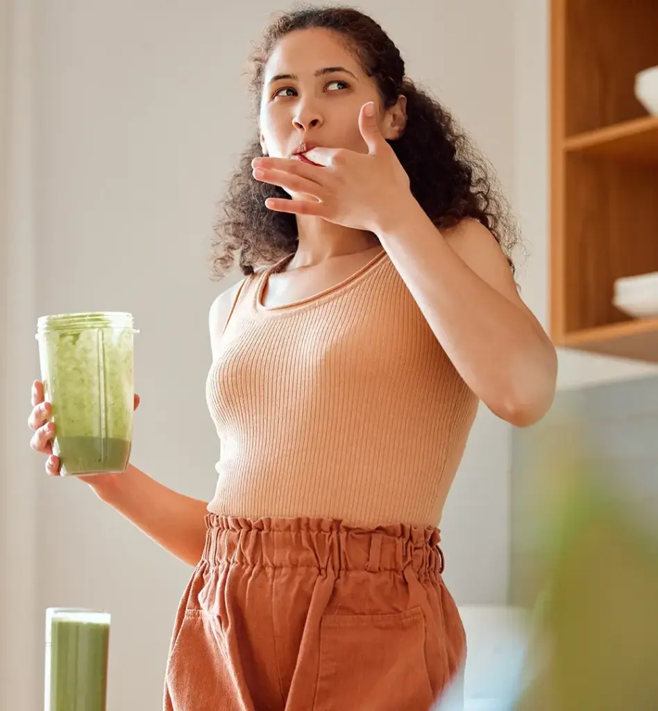 Eine Frau steht in einer Küche. Sie lutscht sich den Zeigefinger ihrer linken Hand ab. In der rechten Hand hält sie einen Behälter für einen Mixer, in dem ein grüner Smoothie drin ist. Das Bild wird im Bereich "myWEIGHTcoach und myHEALTHcoach" verwendet.