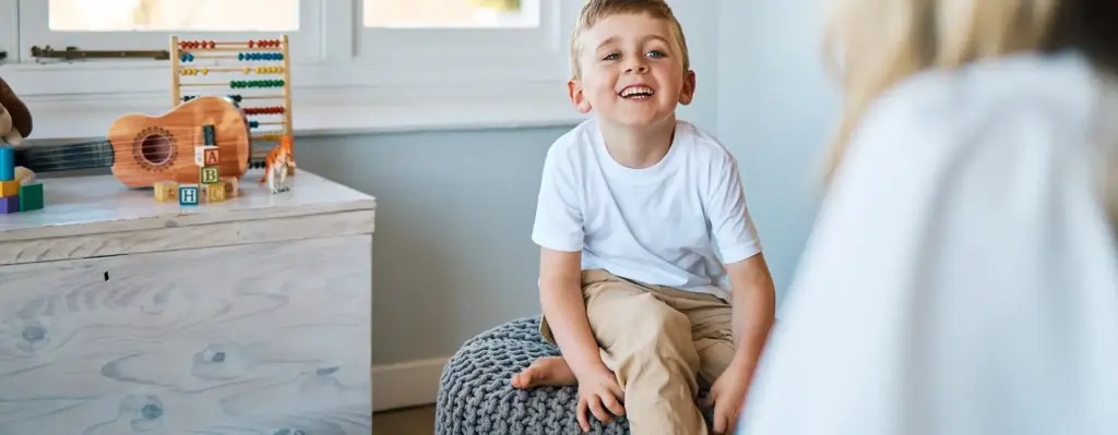 Das Bild zeigt einen Jungen, der auf einem Sitzhocker in einem Kinderzimmer sitzt und lächelt. Das Bild wird im Bereich "Neolexon logopädische Unterstützung für Kinder" verwendet.