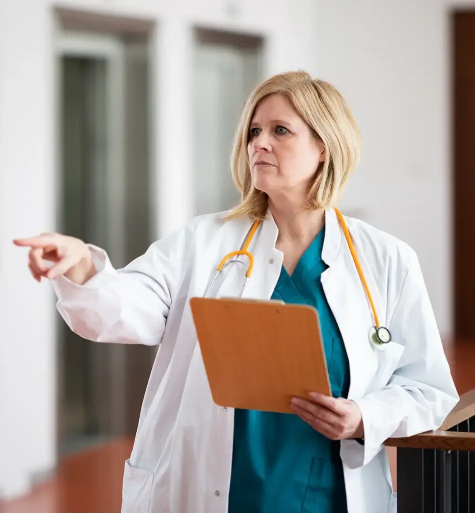 Das Bild zeigt eine Frau im weißen Kittel mit Stethoskop um den Hals gelegt. Sie hat ein Klemmbrett in der Hand und zeigt mit ihrem rechten Zeigefinger auf etwas außerhalb des Bildes. Das Bild wird im Bereich "Ombudspersonen des Medizinischen Dienstes" verwendet.
