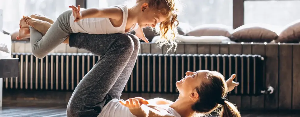 Das Bild zeigt eine Mutter, die mit ihrer jungen Tochter Flieger spielt. Dabei liegt die Mutter auf dem Rücken und balanciert ihre Tochter auf ihren Beinen. Das Bild wird im Bereich "Online Coach Fitness" verwendet.