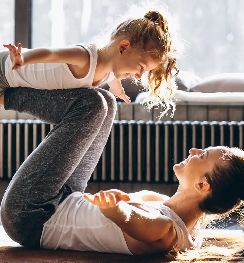 Das Bild zeigt eine Mutter, die mit ihrer jungen Tochter Flieger spielt. Dabei liegt die Mutter auf dem Rücken und balanciert ihre Tochter auf ihren Beinen. Das Bild wird im Bereich "Online Coach Fitness" verwendet.