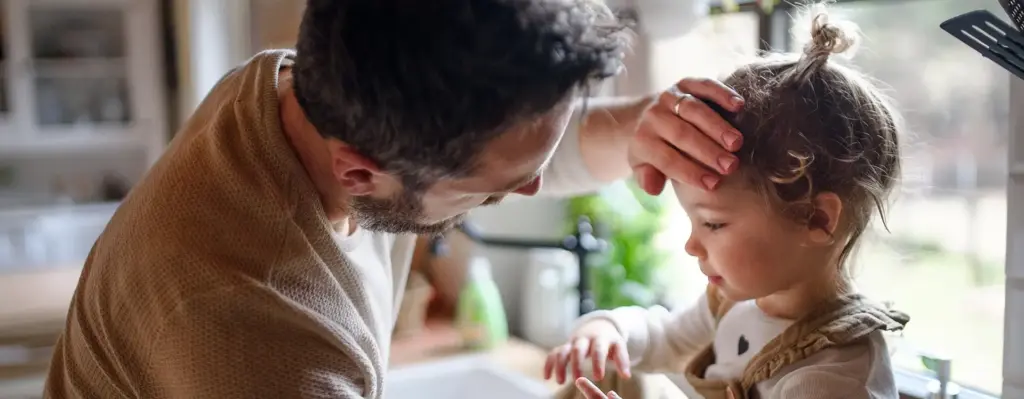 Das Bild zeigt ein kleines Mädchen, dass auf einer Küchenzeile sitzt. Der Vater des Mädchens fühlt mit der linken Hand die Stirn des Mädchens als würde er die Temperatur messen und schaut sie leicht gebückt dabei an. Das Bild wird im Bereich "Online Coach Kindergesundheit" verwendet.