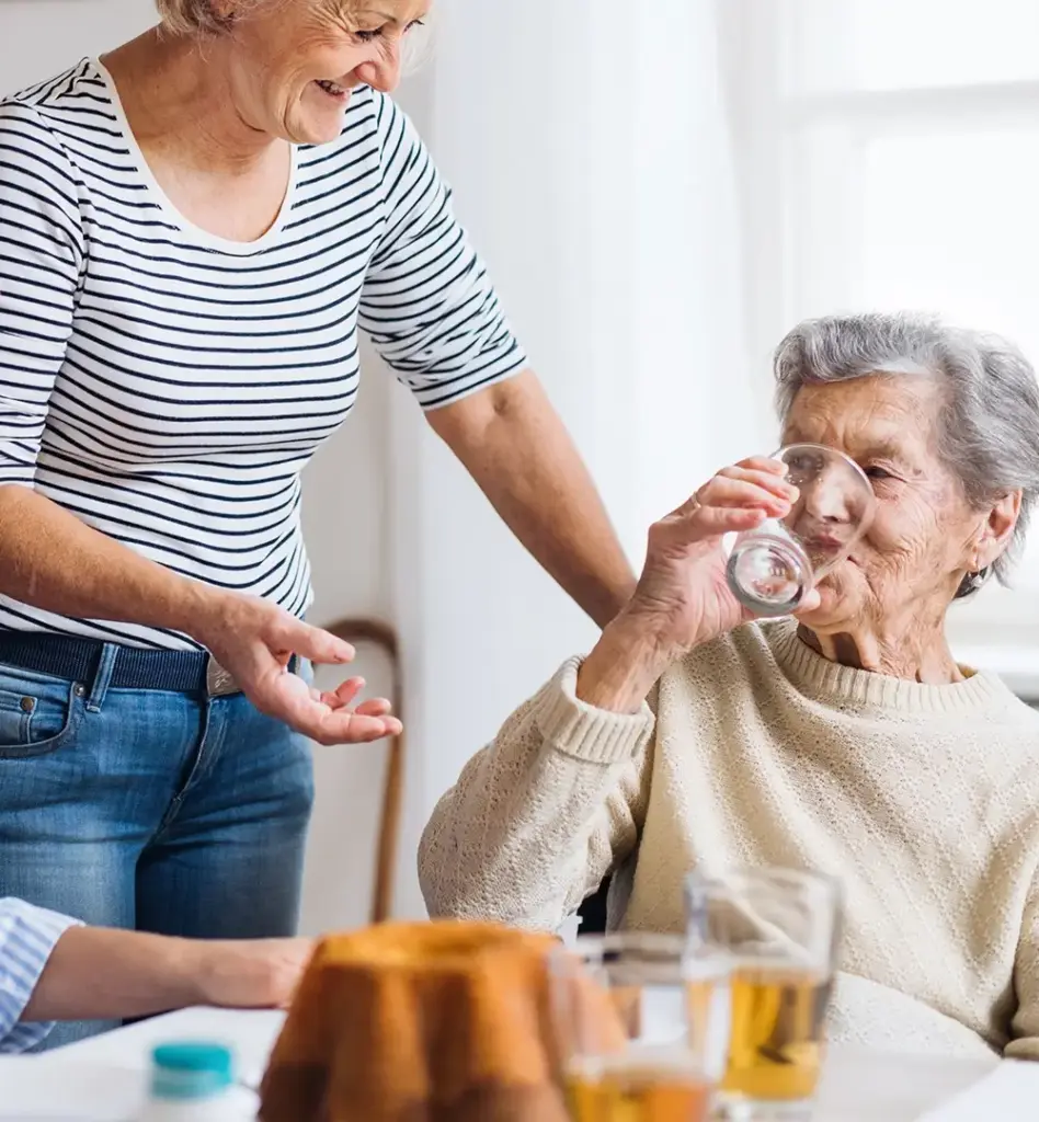 Das Bild zeigt eine ältere Frau im Rollstuhl, die ein Glas Wasser trinkt und an einem Tisch sitzt. Neben ihr steht eine weitere Frau mittleren Alters, die ihre Tochter sein könnte. Am Tisch sitzt zudem eine jüngere Frau, die die ältere Dame anlächelt. Das Bild wird im Bereich "Online Coach Pflege" verwendet.