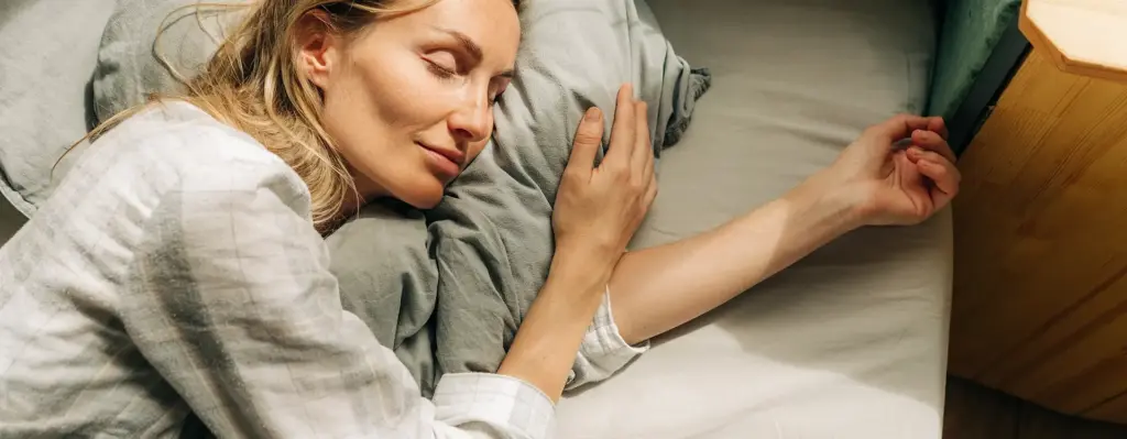 Eine Frau liegt auf ihrer linken Seite im Bett und schläft. Sie hat ein Kissen unter ihrem Kopf und hält es mit der Hand. Sie sieht zufrieden aus. Das Bild wird im Bereich "Online Coach Schlaf" verwendet.