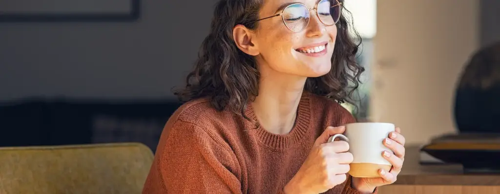 Das Bild zeigt eine Frau, die mit einer Tasse in der Hand auf einer Couch oder einem Sessel sitzt. Sie trägt eine Brille, blickt zufrieden lachend mit geschlossenen Augen und wirkt entspannt. Das Bild wird im Bereich "Online Coach Stress" verwendet.