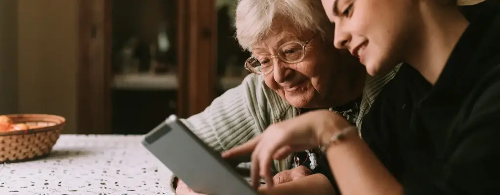 Das Bild zeigt eine ältere Frau zusammen mit einem jüngeren Mädchen, das ihre Enkelin sein könnte. Sie schauen gemeinsam auf ein Tablet und lächeln. Das Bild wird im Bereich "Online Pflegekurse" verwendet.
