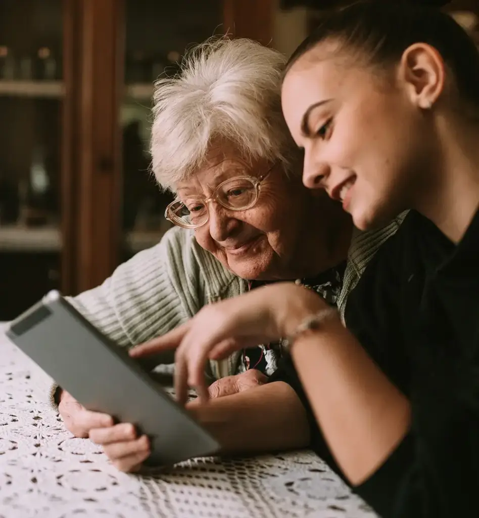 Das Bild zeigt eine ältere Frau zusammen mit einem jüngeren Mädchen, das ihre Enkelin sein könnte. Sie schauen gemeinsam auf ein Tablet und lächeln. Das Bild wird im Bereich "Online Pflegekurse" verwendet.