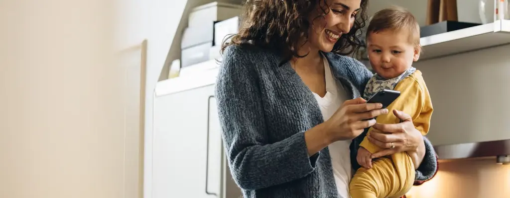 Das Bild zeigt eine Frau mit Kleinkind auf dem Arm. Sie blickt auf ein Smartphone und lächelt. Sie stehen in einer Küche. Das Bild wird im Bereich "Online-Service „Meine energie-BKK“ App" verwendet.