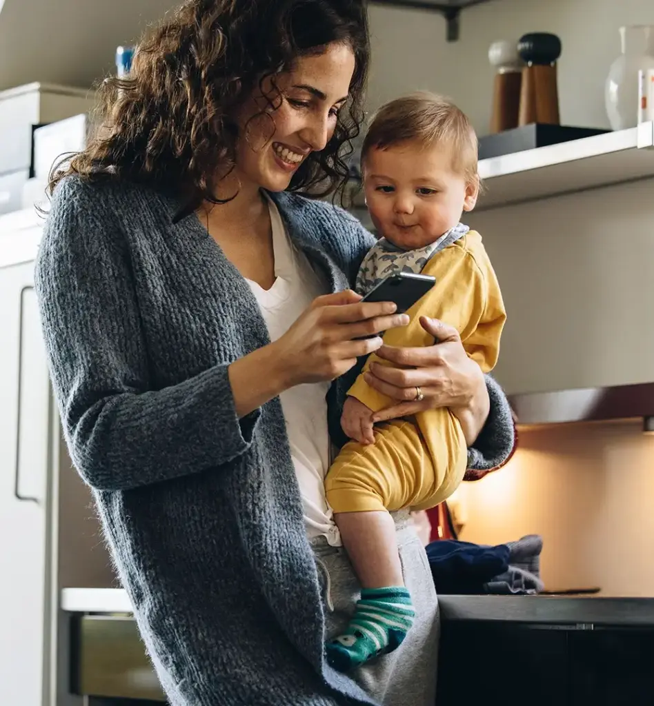 Das Bild zeigt eine Frau mit Kleinkind auf dem Arm. Sie blickt auf ein Smartphone und lächelt. Sie stehen in einer Küche. Das Bild wird im Bereich "Online-Service „Meine energie-BKK“ App" verwendet.