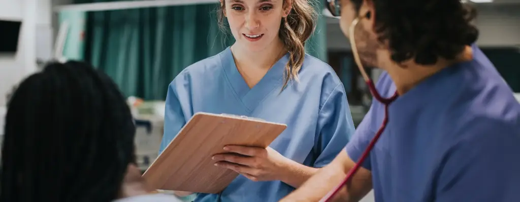 Das Bild zeigt eine im Krankenhaus arbeitende Frau im blauen Kittel. Sie hält ein Klemmbrett in der Hand. Unscharf im Vordergrund sieht man einen männlichen Kollegen und einen Patient oder eine Patientin. Das Bild wird im Bereich "Patientenrechte" verwendet.