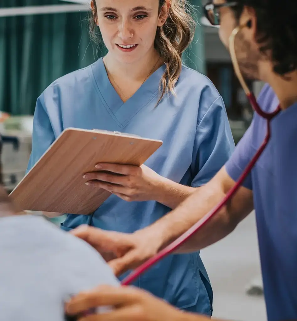 Das Bild zeigt eine im Krankenhaus arbeitende Frau im blauen Kittel. Sie hält ein Klemmbrett in der Hand. Unscharf im Vordergrund sieht man einen männlichen Kollegen und einen Patient oder eine Patientin. Das Bild wird im Bereich "Patientenrechte" verwendet.