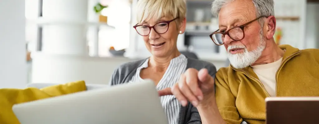 Ein Mann und eine Frau zwischen 60 und 70 Jahren alt sitzen auf einem Sofa. Die Frau hält einen Laptop auf dem Schoß und blickt auf den Bildschirm. Der Mann zeigt mit der Hand auf den Bildschirm und blickt ebenfalls dort hin. Das Bild wird im Bereich "Pflegefinder" verwendet.