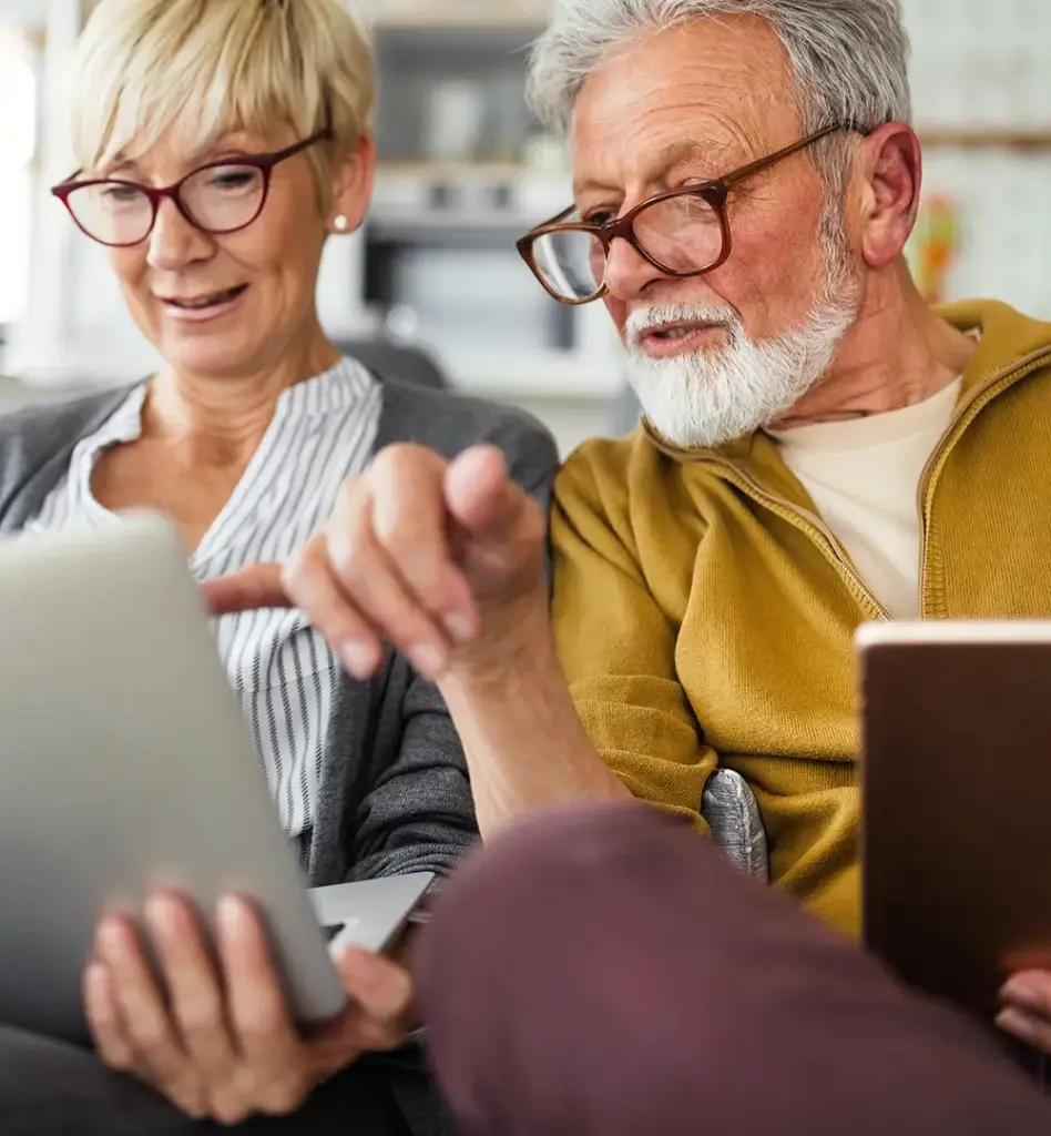 Ein Mann und eine Frau zwischen 60 und 70 Jahren alt sitzen auf einem Sofa. Die Frau hält einen Laptop auf dem Schoß und blickt auf den Bildschirm. Der Mann zeigt mit der Hand auf den Bildschirm und blickt ebenfalls dort hin. Das Bild wird im Bereich "Pflegefinder" verwendet.