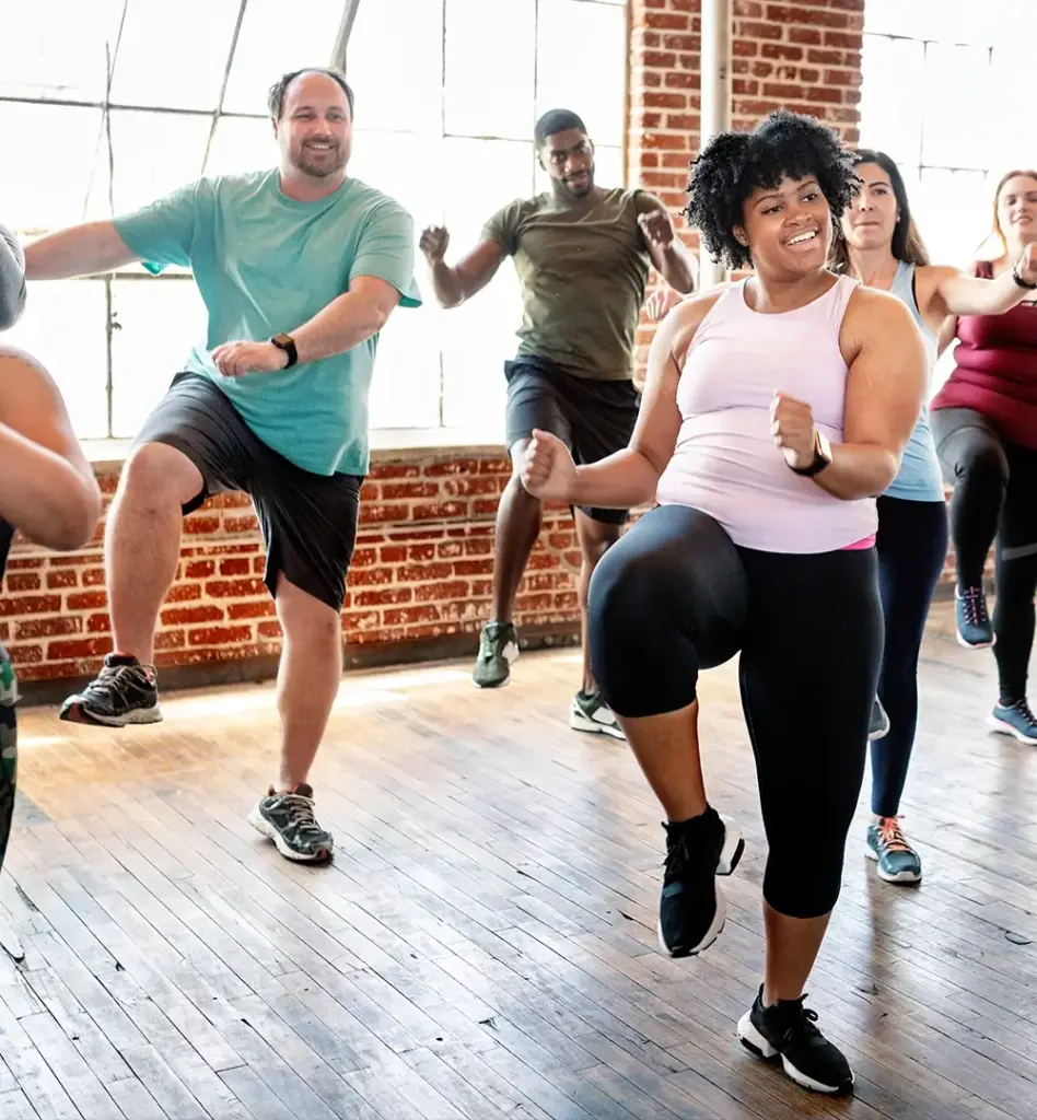 Das Bild zeigt eine Gruppe von sechs Personen, die gemeinsam in einem Raum Sport machen. Dabei machen sie alle die gleiche Bewegung, in dem sie ein Bein seitlich hochheben und die Arme dazu nehmen. Das Bild wird im Bereich "Präventionskurse" verwendet.