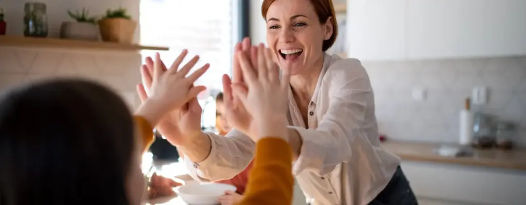 Das Bild zeigt eine Frau mit kurzen rötlichen Haaren. Sie steht in einer Küche und beugt sich mit beiden Händen nach vorne zu einem Kind. Kind und Frauen geben sich ein doppeltes High Five. Das Bild wird im Bereich "Private Zusatzversicherung ExtraPlus" verwendet.