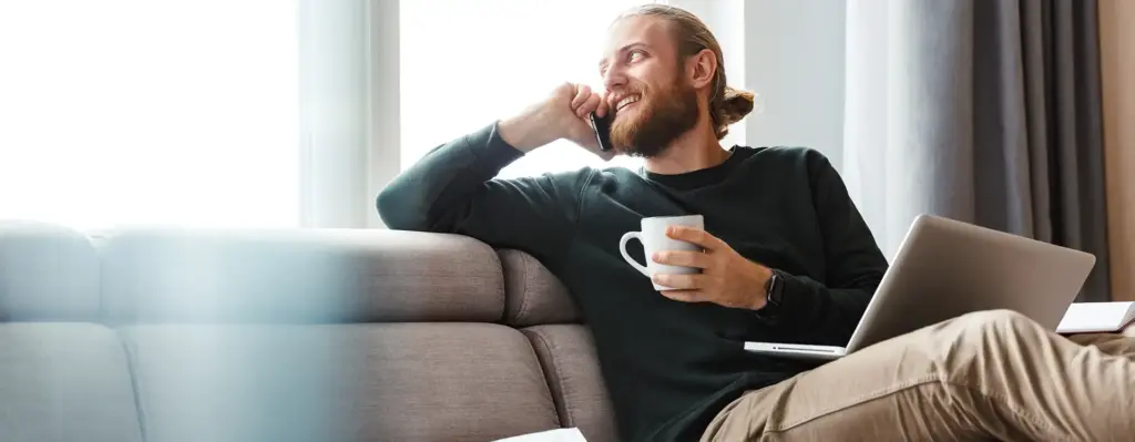 Ein Mann sitzt auf der Couch und hält in der linken Hand eine Tasse. Mit dem rechten Arm lehnt er auf der Sofalehne und telefoniert mit einem Smartphone. Auf seinem Schoß steht ein Laptop. Das Bild wird im Bereich "Psychische Gesundheit am Arbeitsplatz" verwendet.