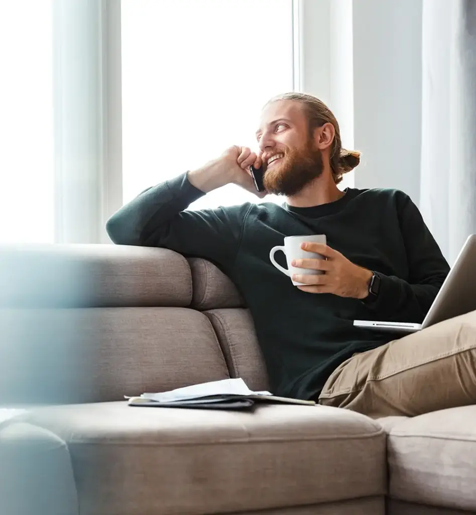 Ein Mann sitzt auf der Couch und hält in der linken Hand eine Tasse. Mit dem rechten Arm lehnt er auf der Sofalehne und telefoniert mit einem Smartphone. Auf seinem Schoß steht ein Laptop. Das Bild wird im Bereich "Psychische Gesundheit am Arbeitsplatz" verwendet.
