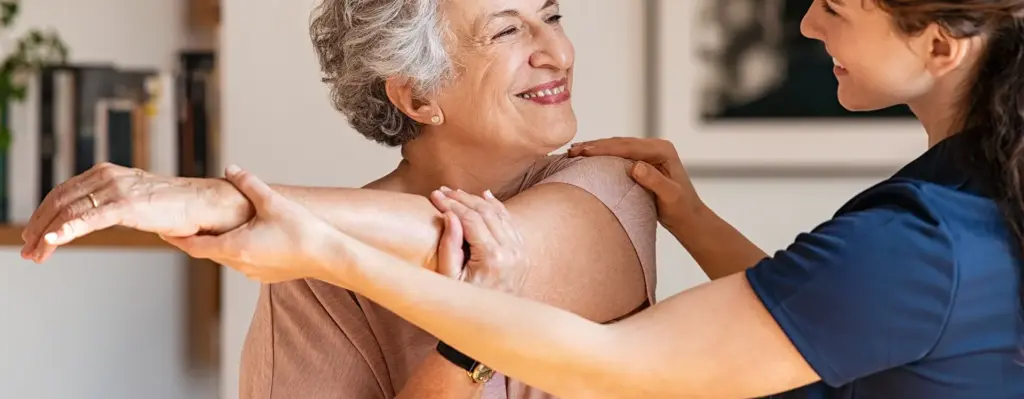 Eine ältere Frau dehnt ihren Arm. Eine jüngere Frau unterstützt sie dabei. Das Bild wird im Bereich "Rehasport und Funktionstraining" verwendet.