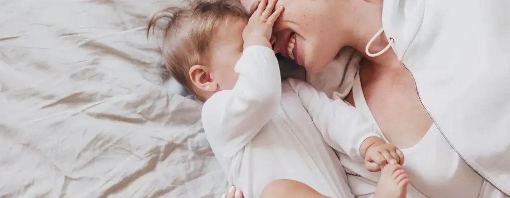 Das Bild zeigt eine Frau mit ihrem Baby. Sie liegen beide auf einem Bett. Das Kind hebt seine Beinchen und fasst der Mutter ins Gesicht. Sie lehnen mit den Köpfen aneinander. Das Bild wird im Bereich "RSV Prophylaxe für Kinder" verwendet.