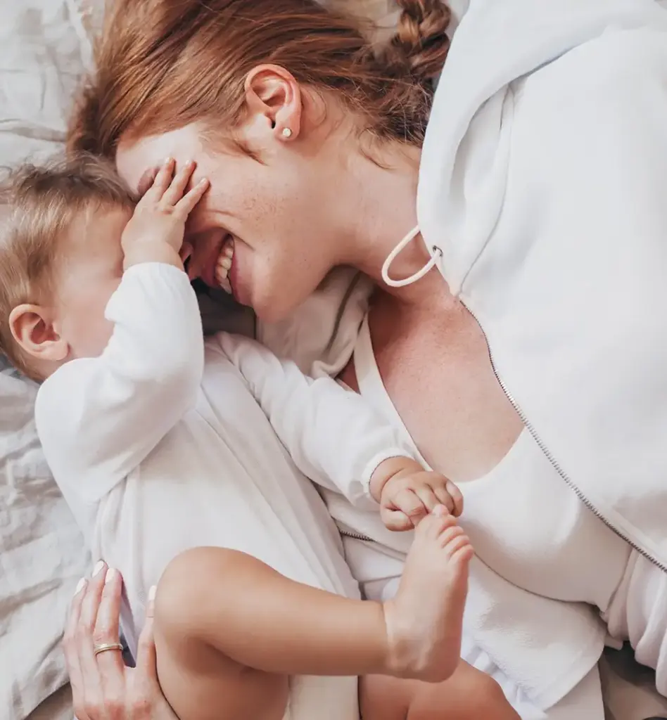 Das Bild zeigt eine Frau mit ihrem Baby. Sie liegen beide auf einem Bett. Das Kind hebt seine Beinchen und fasst der Mutter ins Gesicht. Sie lehnen mit den Köpfen aneinander. Das Bild wird im Bereich "RSV Prophylaxe für Kinder" verwendet.