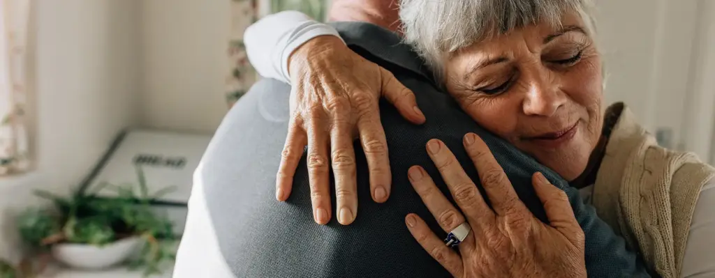 Das Bild zeigt eine ältere Frau, die mit geschlossenen Augen und leichtem Lächeln einen älteren Mann umarmt. Das Bild wird im Bereich "Rythmusleben Schlaganfall Prevention" verwendet.