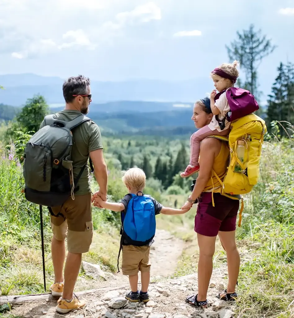 Eine vierköpfige bestehend aus Vater, Mutter, Sohn und Tochter mit Wanderausrüstung steht auf einem Wanderweg. Der Sohn hält die Hande der Eltern, die Tochter sitzt auf den Schultern der Mutter. Beide Eltern schauen sich an. Das Bild wird im Bereich "Schutzimpfung" verwendet.