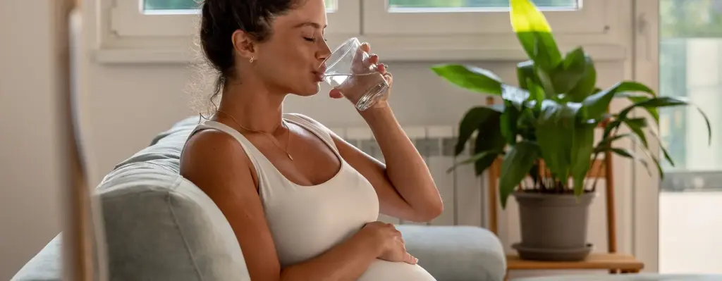 Eine schwangere Frau sitzt auf einem Sofa, hält sich den Bauch und trink mit geschlossenen Augen ein Glas Wasser. Das Bild wird im Bereich "Schwangerschaft und Entbindung" verwendet.