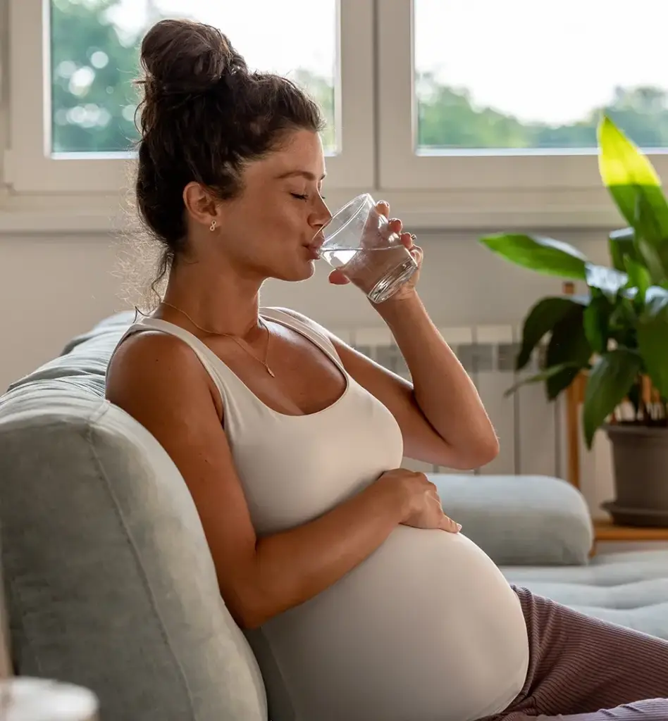 Eine schwangere Frau sitzt auf einem Sofa, hält sich den Bauch und trink mit geschlossenen Augen ein Glas Wasser. Das Bild wird im Bereich "Schwangerschaft und Entbindung" verwendet.