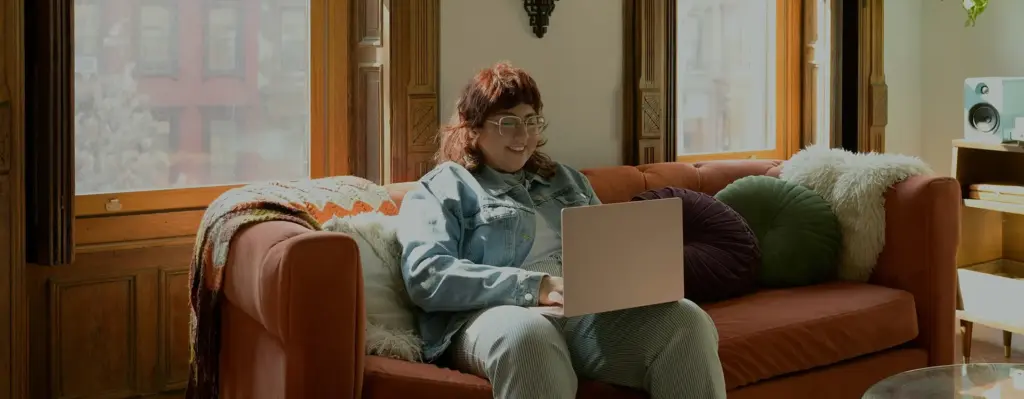 Das Bild zeigt eine junge Frau mit Brille, die auf einem Sofa vor einem Fenster sitzt und auf einen laptop blickt, der auf ihrem Bein steht. Das Bild wird im Bereich "TeleClinic App – 24/7 Online-Sprechstunde" verwendet.