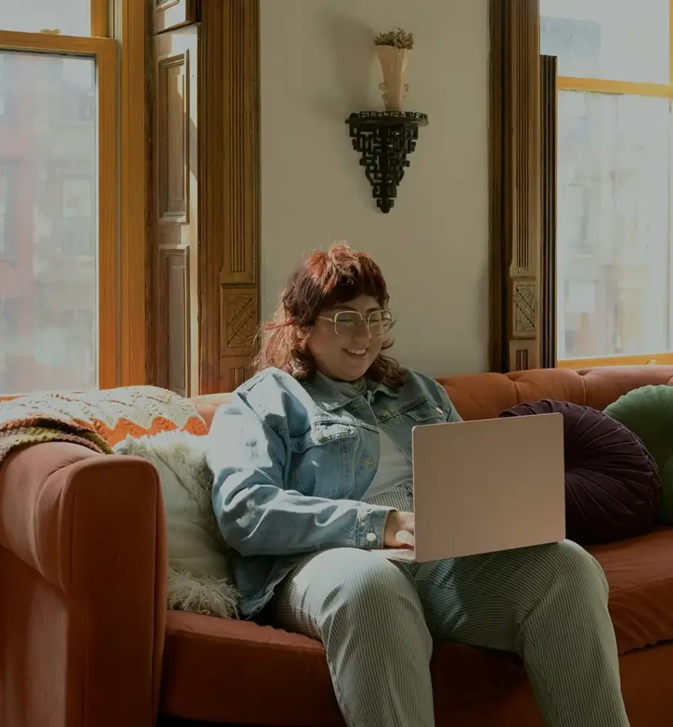 Das Bild zeigt eine junge Frau mit Brille, die auf einem Sofa vor einem Fenster sitzt und auf einen laptop blickt, der auf ihrem Bein steht. Das Bild wird im Bereich "TeleClinic App – 24/7 Online-Sprechstunde" verwendet.