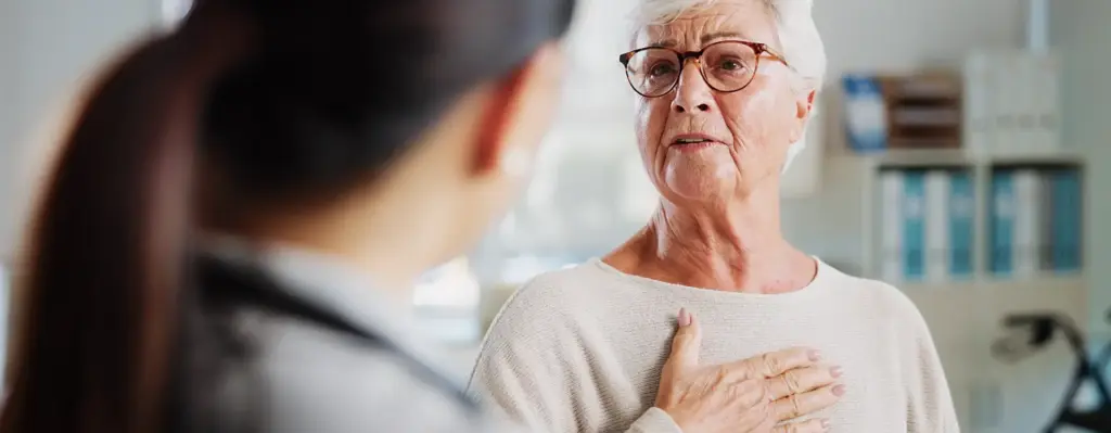 Eine ältere Frau steht einer Ärztin gegenüber. Sie fasst sich mit der rechten Hand auf die Brust, ans Herz. Das Bild wird im Bereich "BKK TeleCoach COPD" verwendet.