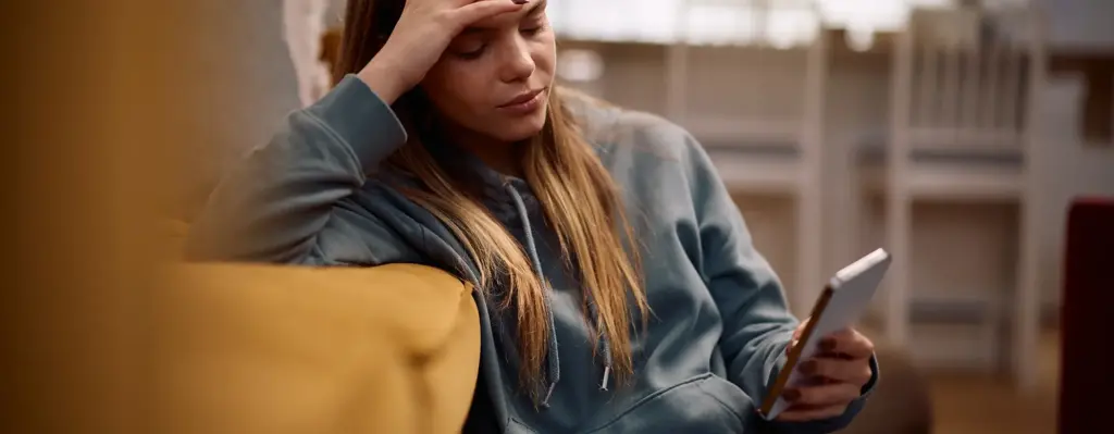 Das Bild zeigt eine junge Frau, die vor einer Couch auf dem Boden sitzt. Sie schaut auf ein Smartphone in ihrer linken Hand. Mit der rechten Hand lehnt sie auf dem Sofa und stützt ihren Kopf. Das Bild wird im Bereich "TeleCoach Kopfschmerz" verwendet.