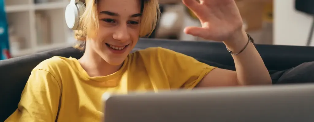 Das Bild zeigt einen Teenager, der auf einer Couch sitzt, Kopfhörer trägt und den Laptop auf seinen Knien hat. Er macht eine grüßende Handbewegung in Richtung Screen. Das Bild wird im Bereich "Videobasierte Online-Psychotherapie bei Kindern und Jugendlichen" verwendet.