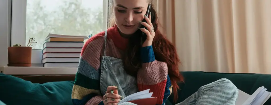 Das Bild zeigt eine junges erwachsenes Mädchen in Pull und Latzhose. Das Mädchen sitzt im Schneidersitz auf einer Couch, hat ein Heft auf ihrem einen Bein liegen und telefoniert mit dem Smartphone. Das Bild wird im Bereich "ViDiKi – Telemedizinische Beratung für Kinder und Jugendliche mit Diabetes Typ 1" verwendet.