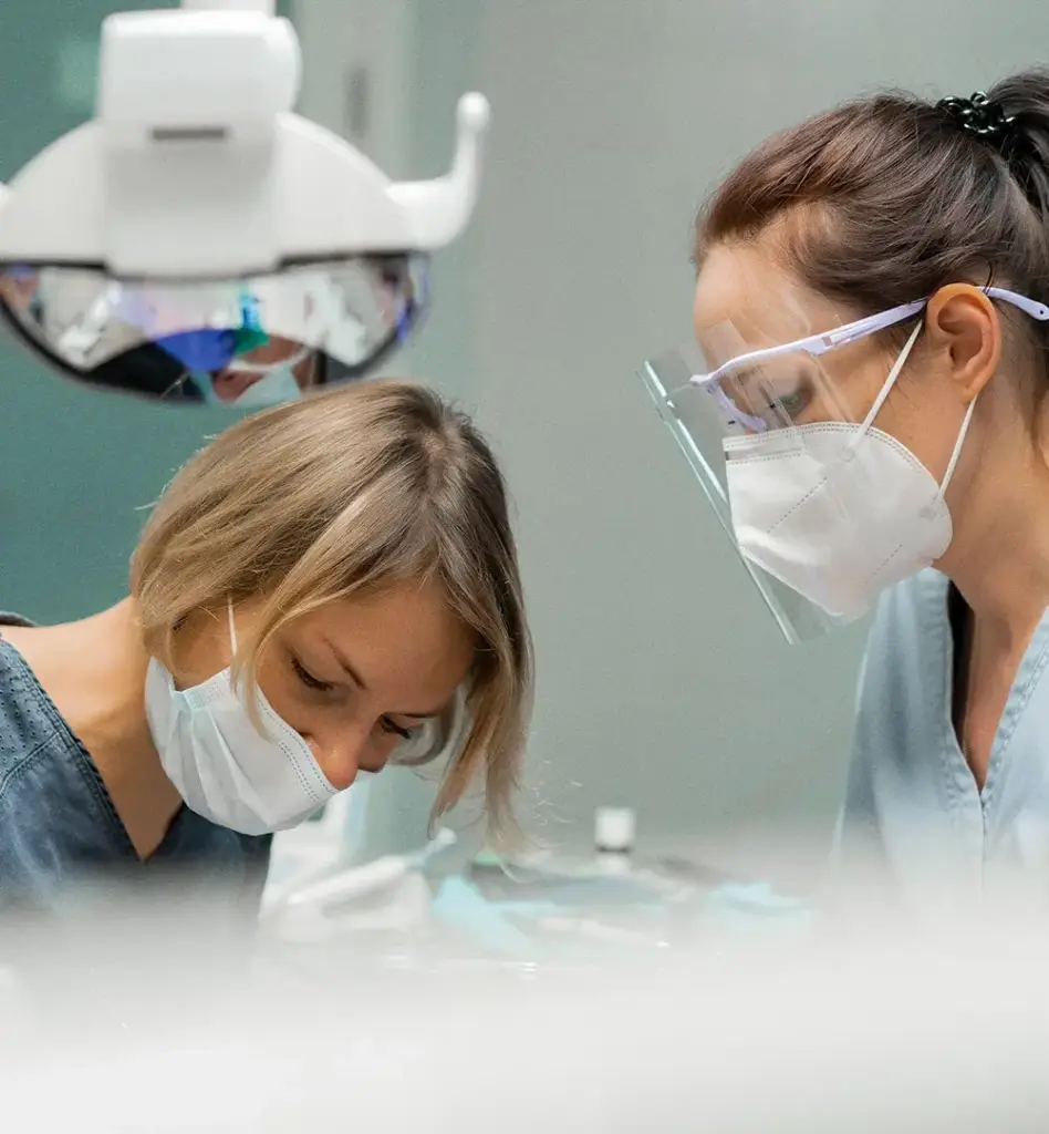 Das Bild zeigt zwei beim Zahnarzt arbeitende Personen während einer Behandlung. Sie tragen Schutzmasken und eine von ihnen auch eine Schutzbrille. Das Bild wird im Bereich "Vollnarkose und Lachgas-Sedierung bei Entfernung der Weisheitszähne" verwendet.