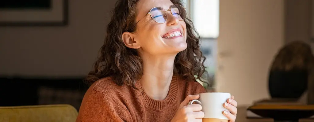 Das Bild zeigt eine Frau, die mit einer Tasse in der Hand auf einer Couch oder einem Sessel sitzt. Sie trägt eine Brille, blickt zufrieden lachend mit geschlossenen Augen leicht nach oben. Das Bild wird im Bereich "Wahltarif PrämiePlus - Mit PrämiePlus bis zu 400 Euro Cashback jährlich – Ihre Gesundheit zahlt sich aus!" verwendet, um den Vorteil für Versicherte und die damit einhergehende Zufriedenheit zu unterstreichen.