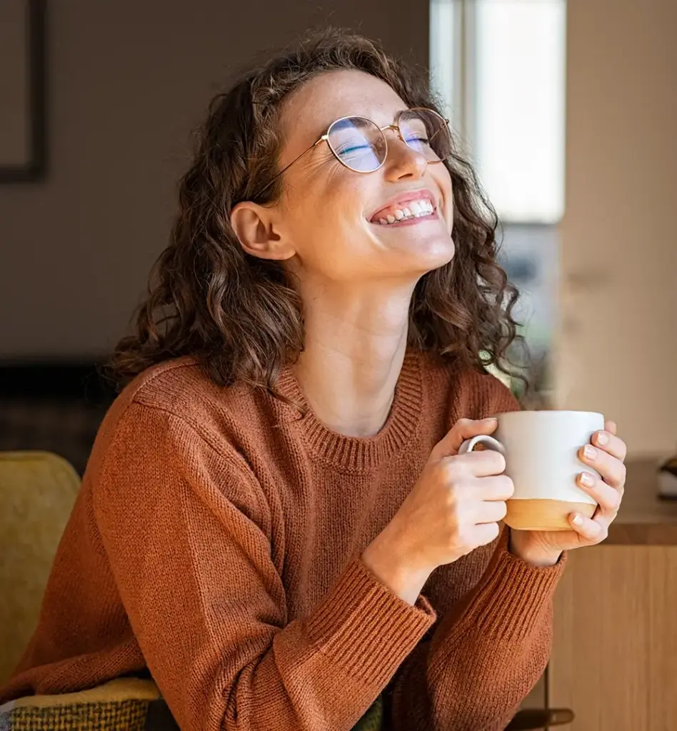 Das Bild zeigt eine Frau, die mit einer Tasse in der Hand auf einer Couch oder einem Sessel sitzt. Sie trägt eine Brille, blickt zufrieden lachend mit geschlossenen Augen leicht nach oben. Das Bild wird im Bereich "Wahltarif PrämiePlus - Mit PrämiePlus bis zu 400 Euro Cashback jährlich – Ihre Gesundheit zahlt sich aus!" verwendet, um den Vorteil für Versicherte und die damit einhergehende Zufriedenheit zu unterstreichen.