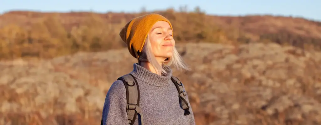 Das Bild zeigt eine Frau, die mit geschlossenen Augen die Sonne genießt. Sie steht mit Wanderstöcken und einem Rucksack auf dem Rücken in der Natur und trägt einen Pullover und eine gelbe Mütze. Das Bild wird im Bereich "Well-Aktiv-Programm" verwendet. 