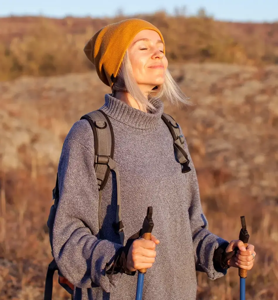 Das Bild zeigt eine Frau, die mit geschlossenen Augen die Sonne genießt. Sie steht mit Wanderstöcken und einem Rucksack auf dem Rücken in der Natur und trägt einen Pullover und eine gelbe Mütze. Das Bild wird im Bereich "Well-Aktiv-Programm" verwendet. 