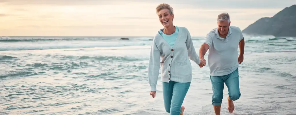 Ein Paar mittleren Alters läuft am Strand durchs Meer und lacht. Die Frau läuft vor und hält dabei die Hand des Mannes. Das Bild wird im Bereich "Wellness und Gesundheitsreisen" verwendet.