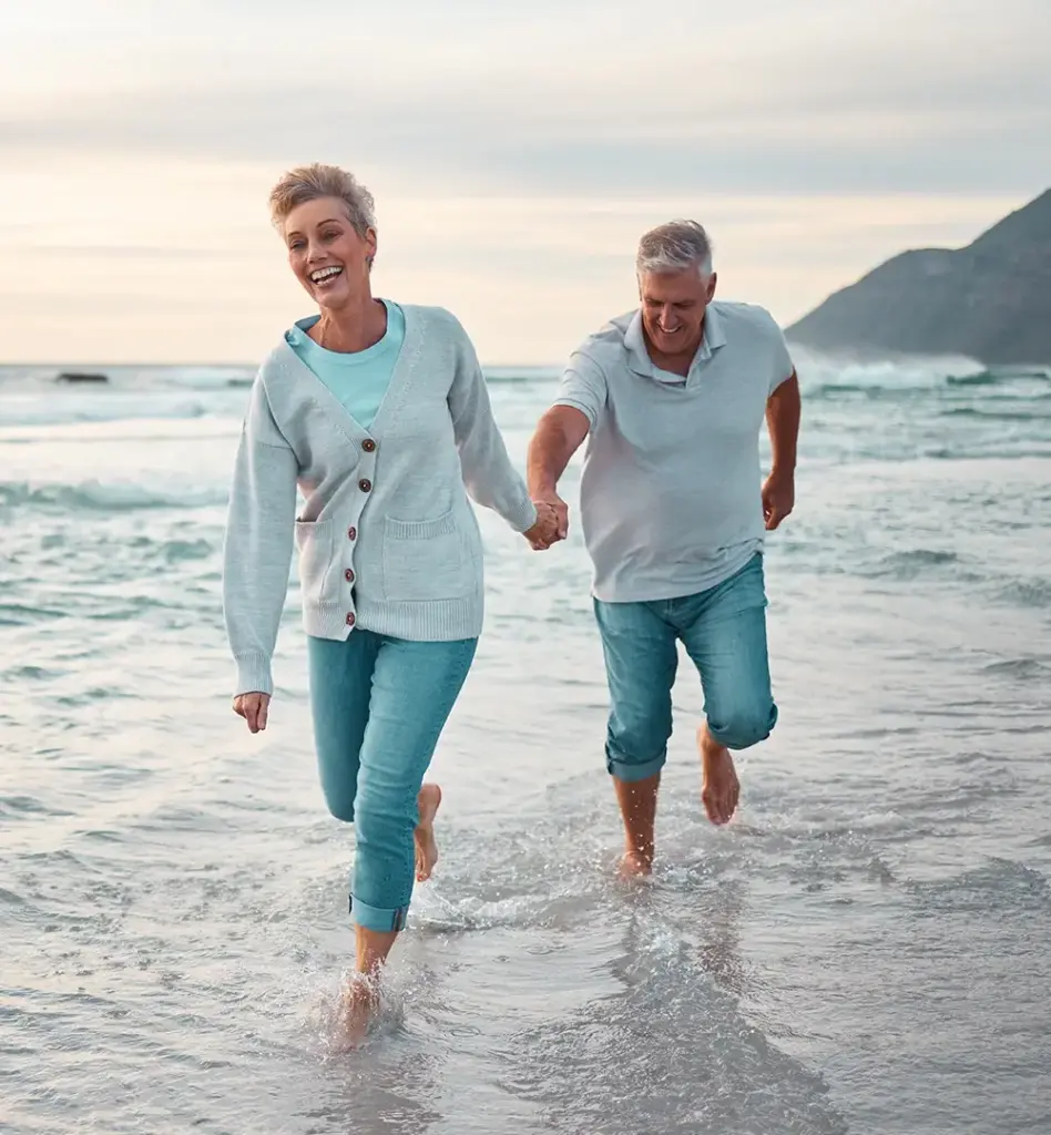 Ein Paar mittleren Alters läuft am Strand durchs Meer und lacht. Die Frau läuft vor und hält dabei die Hand des Mannes. Das Bild wird im Bereich "Wellness und Gesundheitsreisen" verwendet.