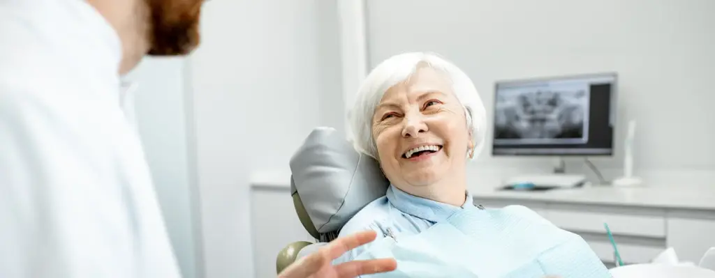 Das Bild zeigt eine ältere grauhaarige Frau, die auf einem Behandlungsstuhl beim Zahnarzt sitzt. Sie lächelt und hört dem Zahnarzt vor, der ihr etwas erzählt. Das Bild wird im Bereich "Zahnersatz - DentNet" verwendet.