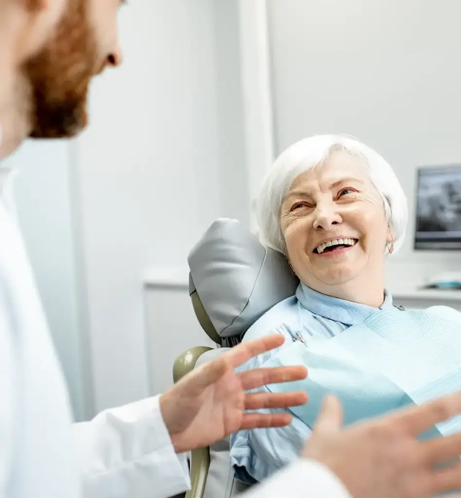 Das Bild zeigt eine ältere grauhaarige Frau, die auf einem Behandlungsstuhl beim Zahnarzt sitzt. Sie lächelt und hört dem Zahnarzt vor, der ihr etwas erzählt. Das Bild wird im Bereich "Zahnersatz - DentNet" verwendet.