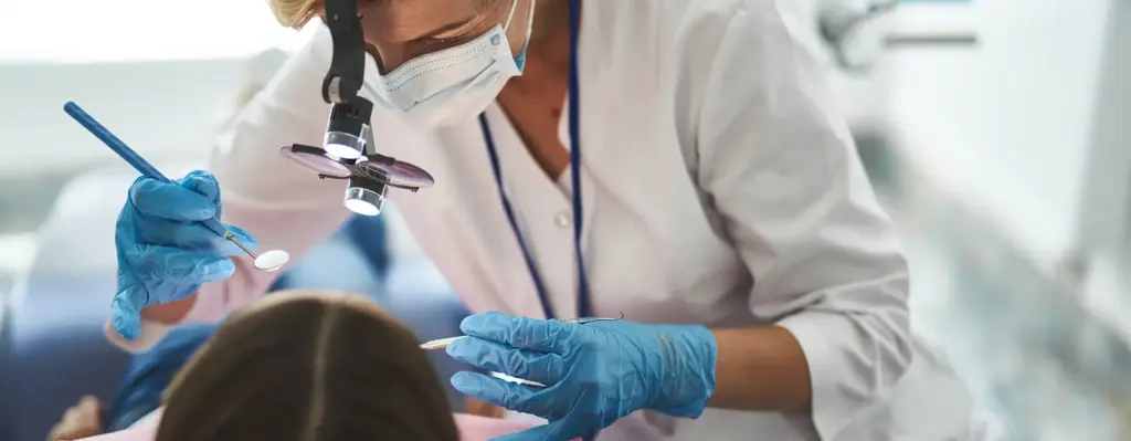Das Bild zeigt eine Zahnärztin, die mit einer Lupenbrille und einem Zahnarztspiegel und weiteren Instrument in den Mund einer Patientin guckt. Die Zahnärztin trägt auch einen Mundschutz und Handschuhe. Das Bild wird im Bereich "Zahnreinigung (PZR)" verwendet.